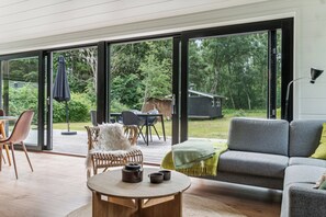 A modern living room with a large sliding glass door leading to a deck, a sofa, a table with tea set, and a view of a forested area.