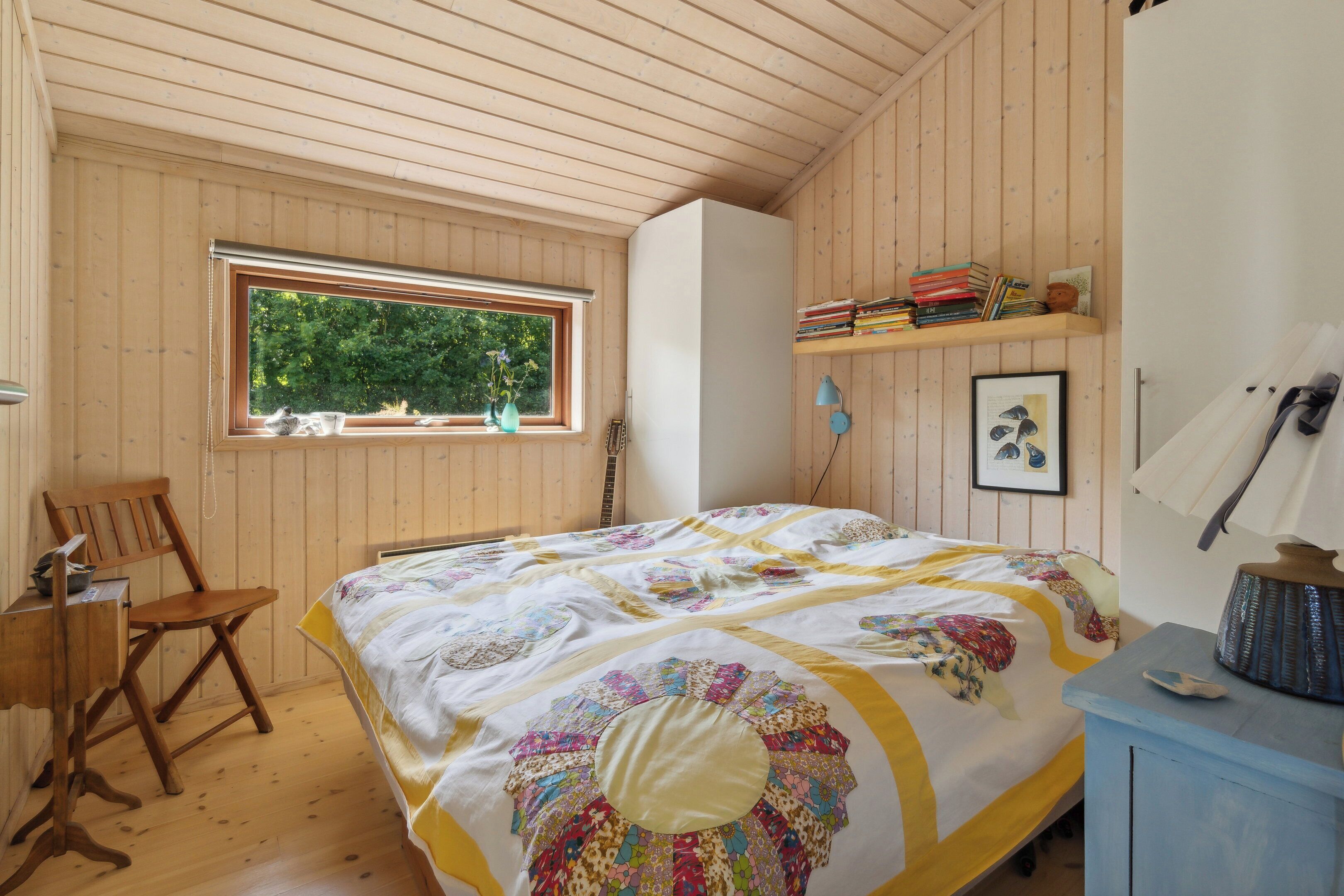 A wooden-paneled room with a bed, a desk, a chair, a window, and a shelf with books and a picture frame.