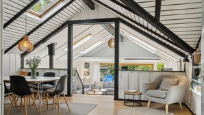 A spacious loft with a high ceiling, exposed beams, a dining area with a table and chairs, a seating area with a sofa, and a kitchen area with a chair and shelves.