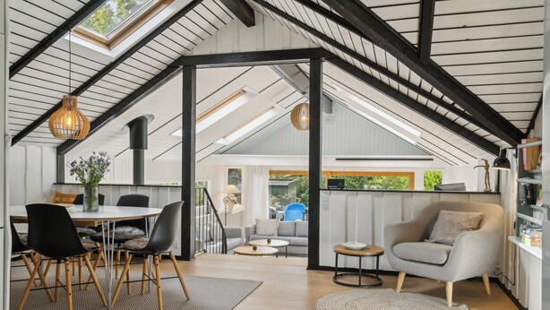 A spacious loft with a high ceiling, exposed beams, a dining area with a table and chairs, a seating area with a sofa, and a kitchen area with a chair and shelves.