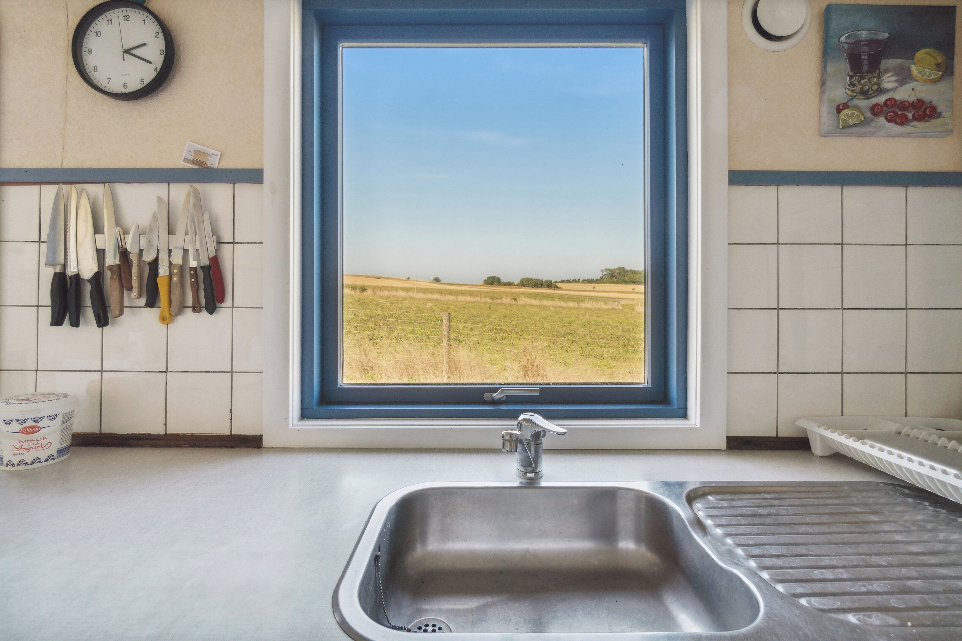 A kitchen with a sink, a window with a view of a field, and a wall-mounted clock.