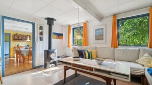 A cozy living room with a sofa, a table, a fireplace, and a view of greenery outside. - Cozy summer house on Bornholm (Nexø)