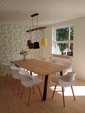 A dining area with a wooden table, white chairs, a floral wallpaper, and a window with a view of greenery.