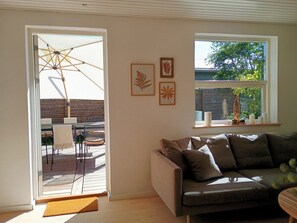 A living room with a sofa, a window with a view of greenery, and a door leading to an outdoor area with a table and chairs.