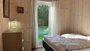 A bedroom with a bed, a bedside table with a lamp, a wooden wardrobe, and a view of a garden through a window with curtains.