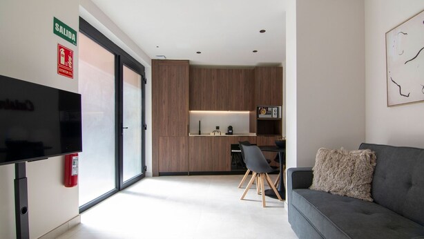 A modern living room with a grey sofa, a flat-screen TV, and a kitchen area with wooden cabinets and a microwave. - VLC HOST - Luz Casanova B (Valencia)