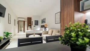 A modern living room with a sofa, a dining table with chairs, and a coffee table. There is a bed in the background, a TV mounted on the wall, and framed artwork on the walls.