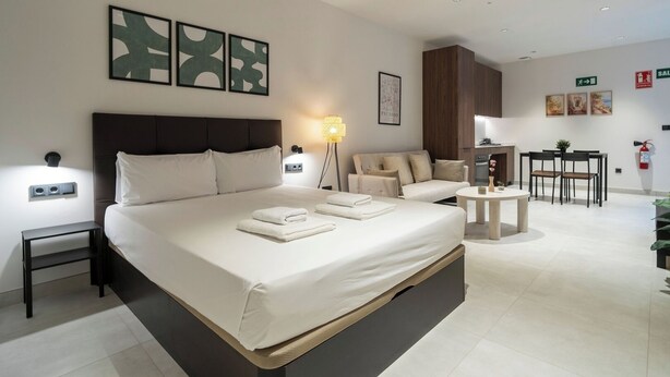 A modern hotel room with a large bed, a sofa, a small table, and a kitchenette.
