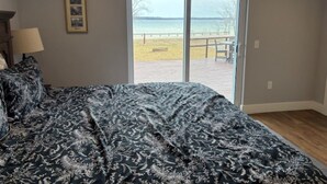 A bedroom with a large bed, a nightstand, a lamp, and a view of a deck and water through a sliding glass door. - Large 6-bedroom home, sleeps 16! WiFi and a private sandy beach in Traverse City (Traverse City)