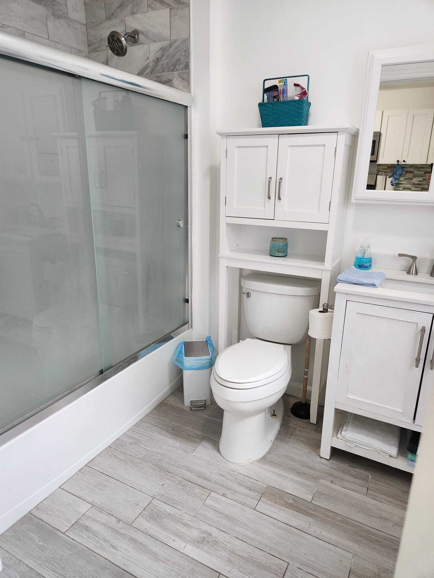 A bathroom with a glass shower enclosure, a toilet, a sink with a mirror, and a small trash bin.