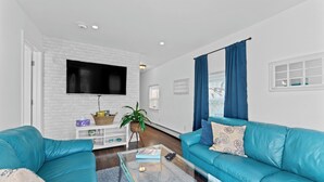 Una sala de estar moderna con una pared de ladrillo blanco, un televisor de pantalla plana montado sobre un mueble blanco, una mesa de centro de vidrio y un sofá azul turquesa con cojines estampados.