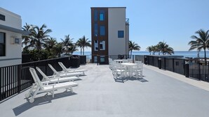 A rooftop terrace with white lounge chairs and tables, a view of the ocean, and a modern building with multiple windows.