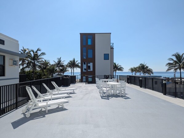 A rooftop terrace with white lounge chairs and tables, a view of the ocean, and a modern building with multiple windows.