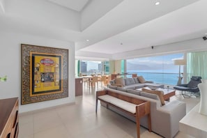 A modern living room with a large sofa, a dining area, and a view of the sea.