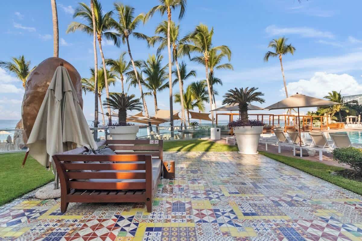 A poolside area with a tiled floor, lounge chairs, umbrellas, and palm trees.