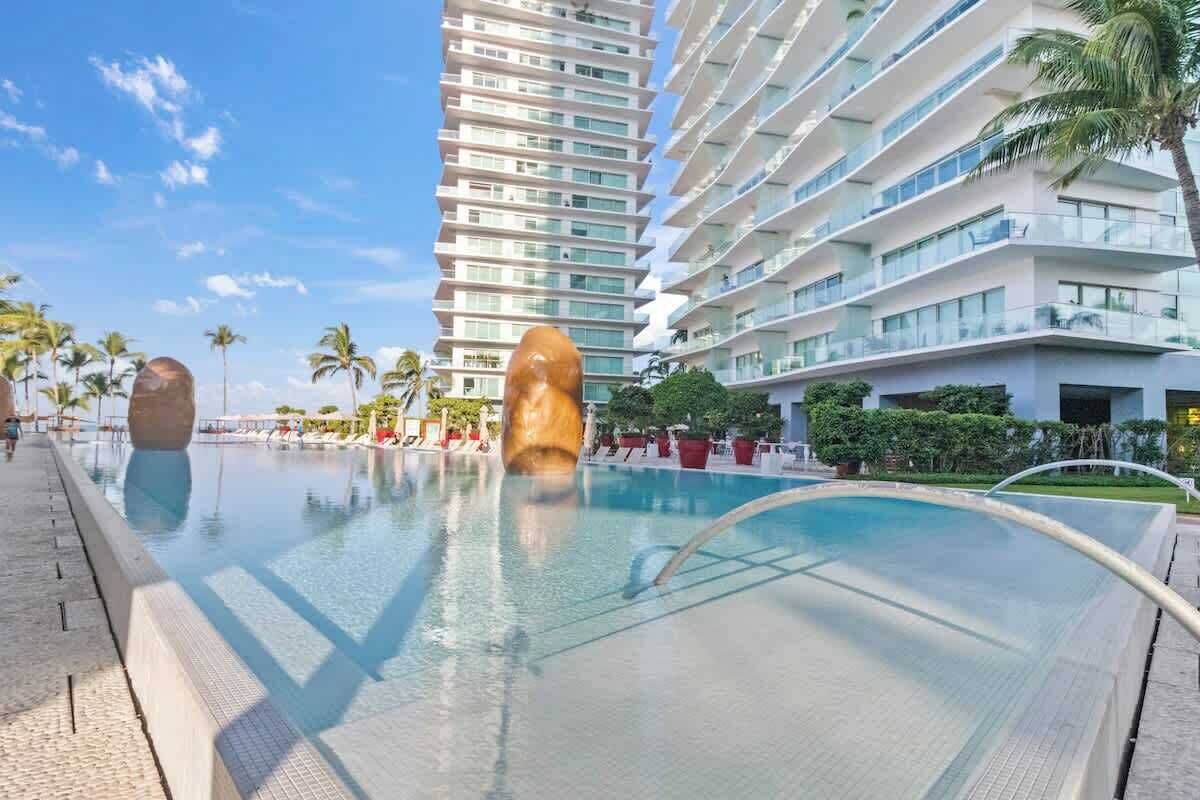 Um hotel moderno com uma piscina grande, palmeiras e um edifício alto ao fundo.