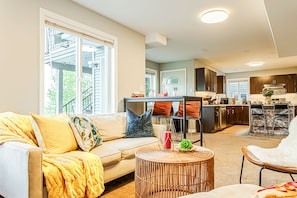 A spacious living room with a sofa, a round wicker coffee table, and a dining area with a kitchen in the background. - 180° Amazing View- Ridge Top, 2 Full Kitchen, 3-Level, Sleeps 15 Comfortably (Chilliwack)