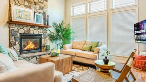 A cozy living room with a stone fireplace, a beige sofa, a coffee table, and a large window with blinds. - 180° Amazing View- Ridge Top, 2 Full Kitchen, 3-Level, Sleeps 16 Comfortably (Chilliwack)