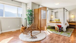 Een slaapkamer met een bed, een houten stoel, een groot raam en uitzicht op bergen.