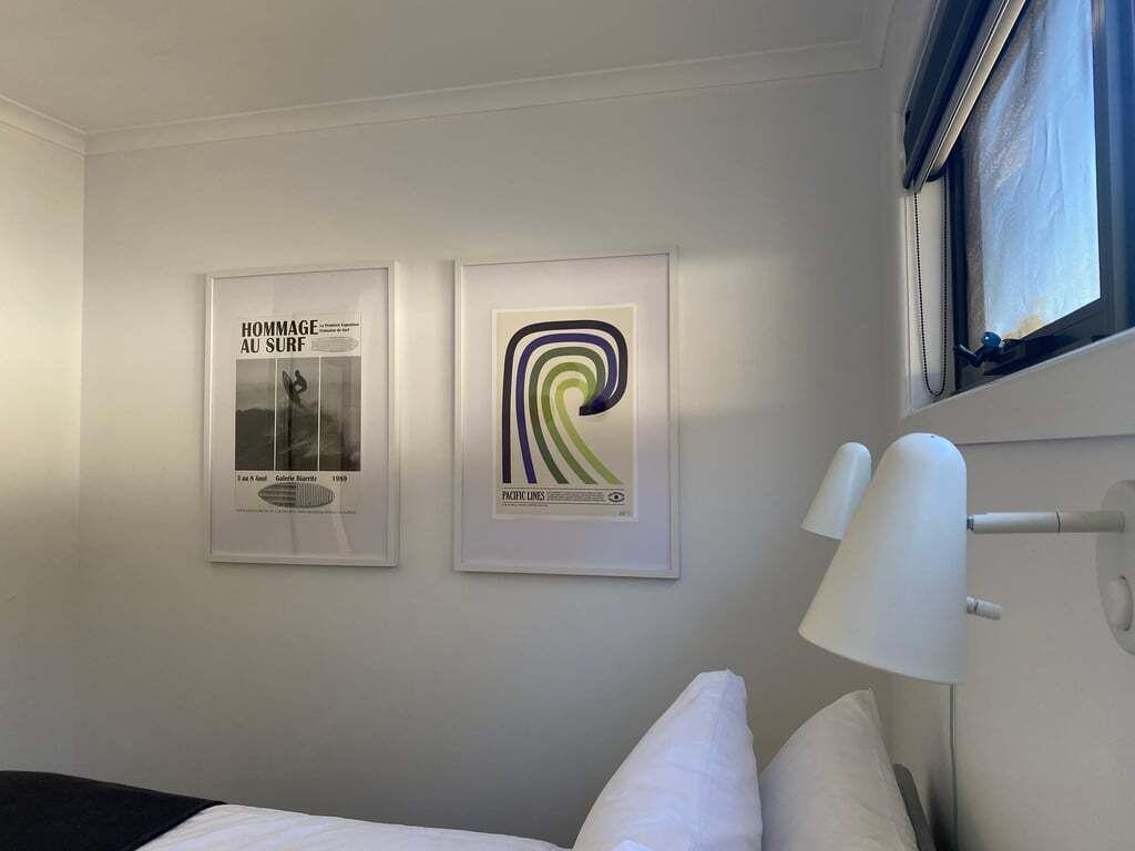 Een hotelkamer met twee ingelijste posters aan de muur, een bed met witte beddengoed en een raam met uitzicht op de lucht.