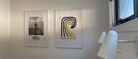 Een hotelkamer met twee ingelijste posters aan de muur, een bed met witte beddengoed en een raam met uitzicht op de lucht.