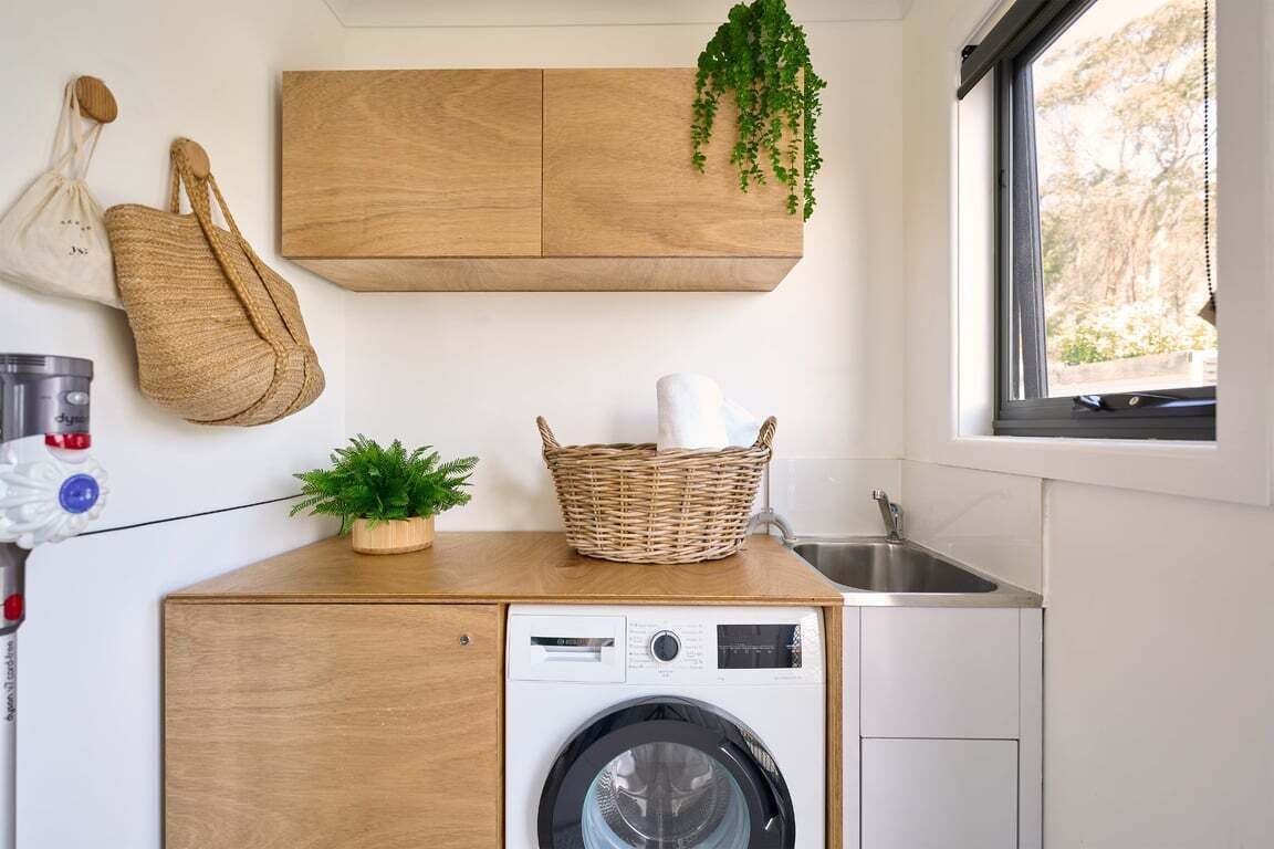 A compact laundry area with a washing machine, a wooden cabinet, a wicker basket, and a hanging plant.