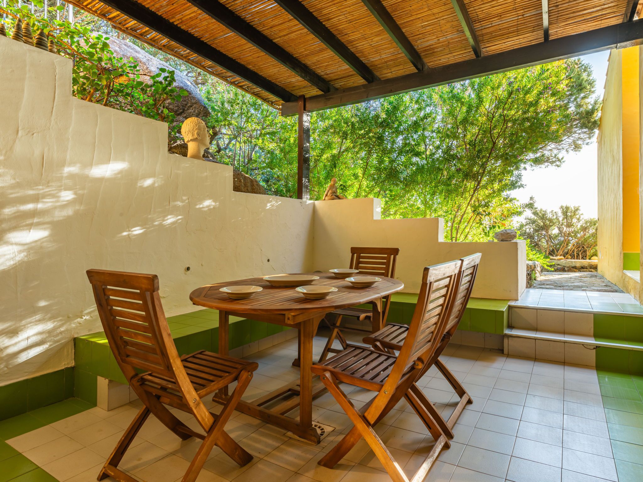 Un espace extérieur abrité, aménagé pour les repas, avec une table ronde et des chaises en bois, entouré d’un mur blanc et d’un sol vert.