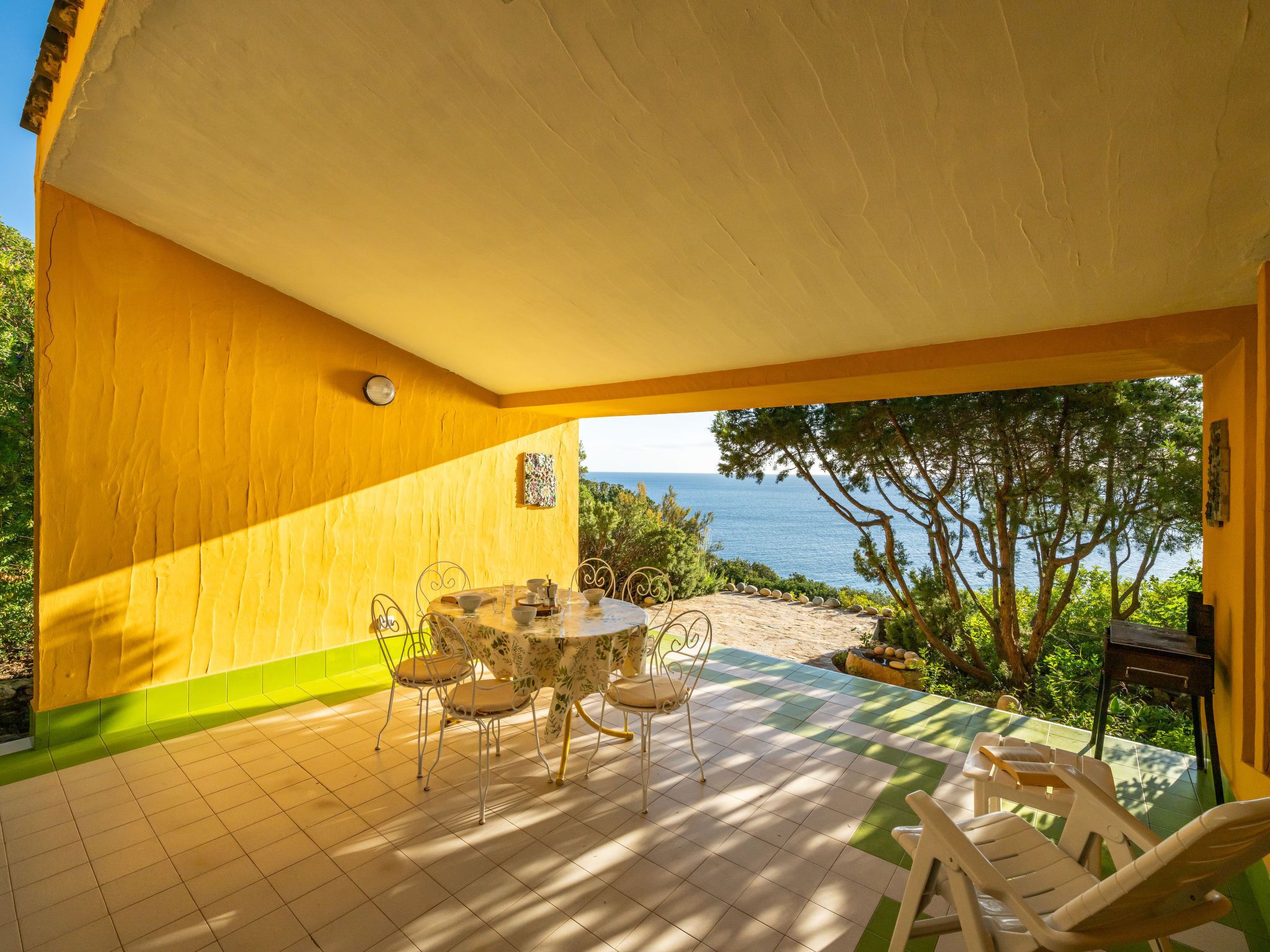 Un patio techado con una mesa y sillas, vista al mar, y un edificio amarillo.