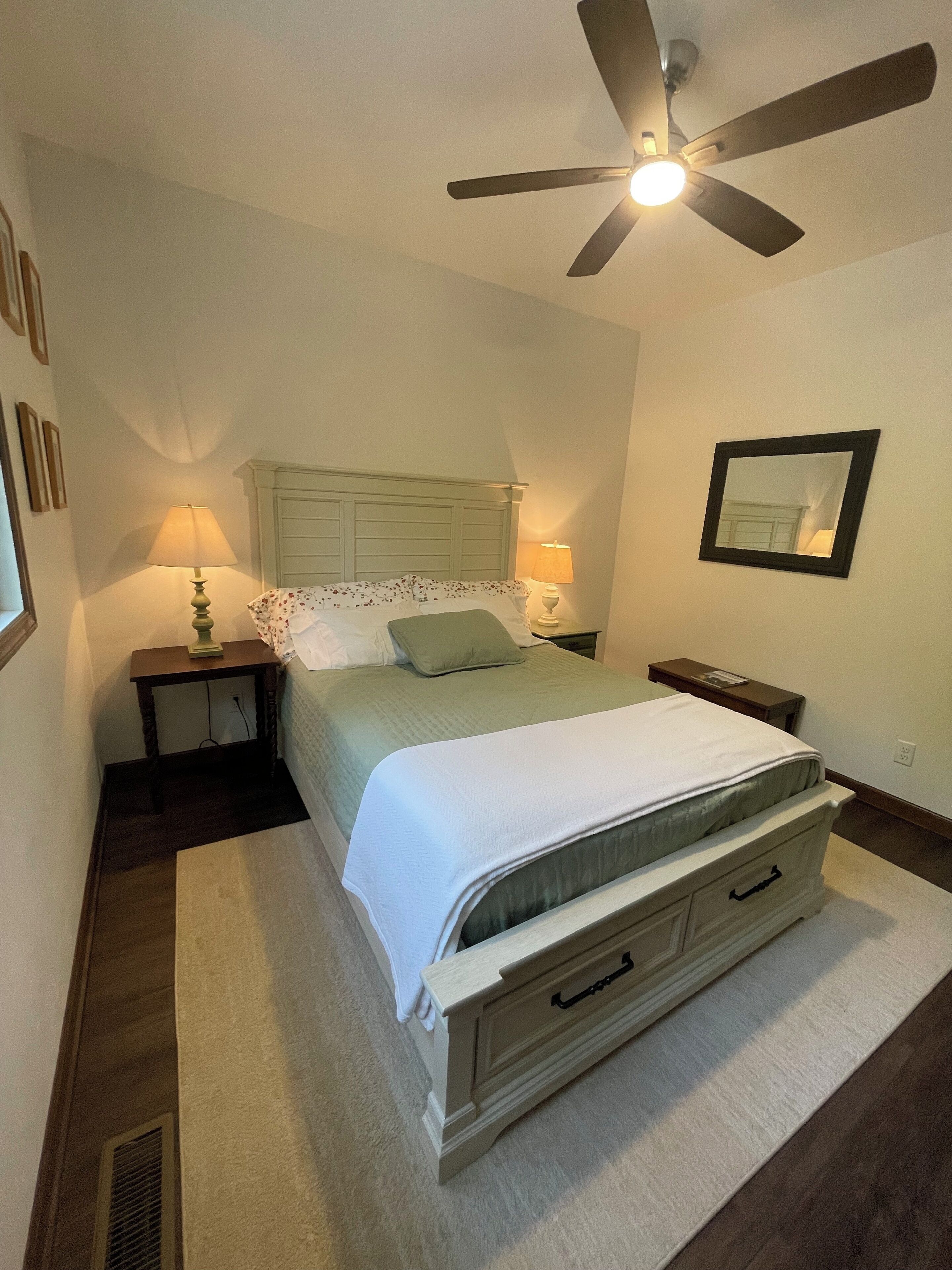 A bedroom with a bed, bedside lamps, a ceiling fan, a nightstand, and a mirror on the wall.