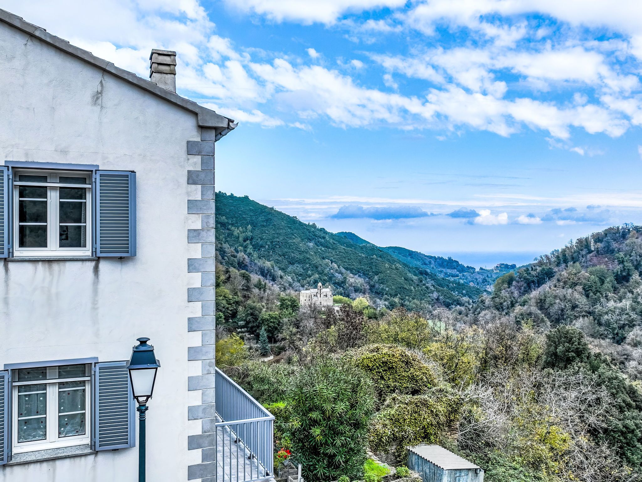 Un edificio blanco con contraventanas azules y un balcón, con vistas a un paisaje montañoso bajo un cielo despejado.