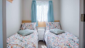 Una habitación pequeña y luminosa con dos camas individuales, cada una con ropa de cama y almohadas a cuadros con motivos florales. Hay una mesita de noche pequeña con una lámpara entre las camas, y una ventana con cortinas azul claro.