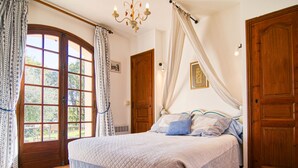 Une chambre à coucher avec un grand lit, un lustre et une vue sur l’extérieur, visible à travers une fenêtre ornée de rideaux.