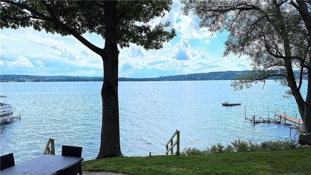 Ein Sitzbereich im Freien mit Tisch und Stühlen, ein Baum und Blick auf einen See mit Steg und Boot.