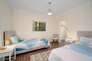 A bedroom with two beds, a nightstand, a chair, a window, and a ceiling light. - Lakeside House Family Escape | Walk to Beach & Lake (Currimundi)