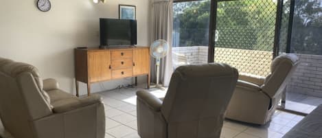 A room with two reclining chairs, a wooden cabinet with a television, and a large window with a view of greenery.