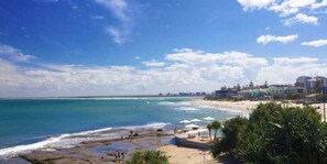 A beach with clear blue water, a sandy shore, and buildings in the background.