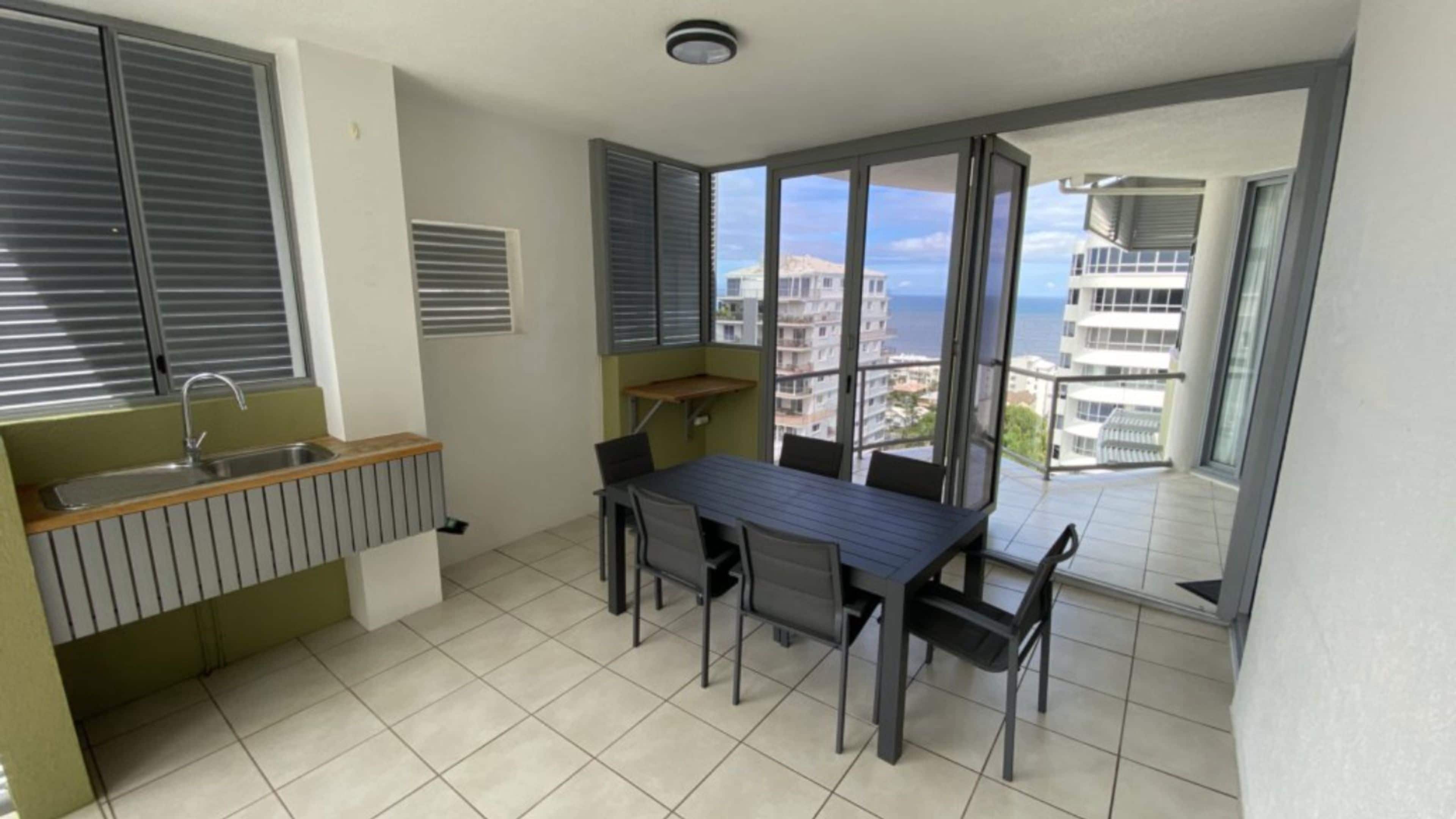 A small kitchenette with a sink, a window with blinds, and a dining area with a table and chairs.