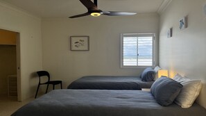 Un dormitorio con dos camas, un ventilador de techo, una ventana con persianas y una pequeña silla.