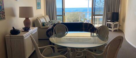 A living room with a glass-top table, wicker chairs, a sofa, a TV, and a view of the ocean.