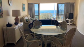 A living room with a glass-top table, wicker chairs, a sofa, a TV, and a view of the ocean.