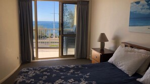 A bedroom with a bed, a nightstand, a lamp, a painting, and a view of the ocean through a sliding door.