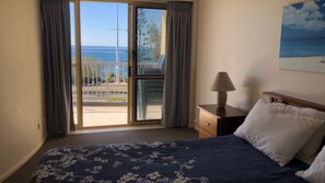 A bedroom with a bed, a nightstand, a lamp, and a view of the ocean through a sliding glass door.
