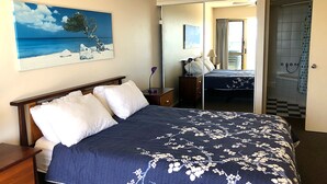 Une chambre à coucher comprenant un lit, une table de chevet, une commode, un miroir et une salle de bain visible à travers une porte ouverte.