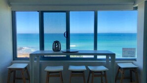 A room with a large window overlooking the ocean, a table with stools, and a small decorative item on the table. - Seaspray U1 Ocean Views & Private Beach (Kings Beach)