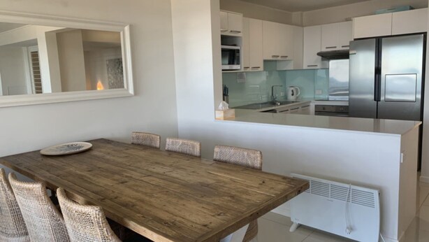 A modern kitchen with stainless steel appliances, a dining area with a wooden table and wicker chairs, and a white radiator. - Seaspray U1 Ocean Views & Private Beach (Kings Beach)