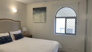 Un dormitorio con una cama, una mesita de noche, una lámpara, un cuadro y una ventana.