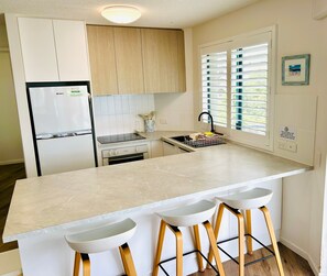 A modern kitchen with a central island, bar stools, a refrigerator, and a window with blinds.
