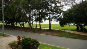 A paved road with a sidewalk on one side, bordered by trees and a grassy field in the background.