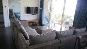 Un espace de vie comprenant un canapé, un téléviseur, une table et un balcon aménagé avec du mobilier d’extérieur.