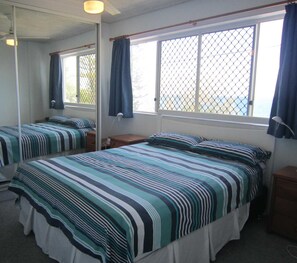 Ein Schlafzimmer mit zwei Betten, einem Fenster mit Vorhängen, einem Nachttisch und einem Spiegel.